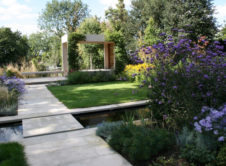 An arts and crafts garden with purple flowers, canal and shelter