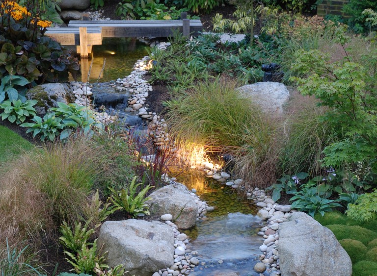 Water feature with wooden bridge and surrounded by stones, grasses and plants