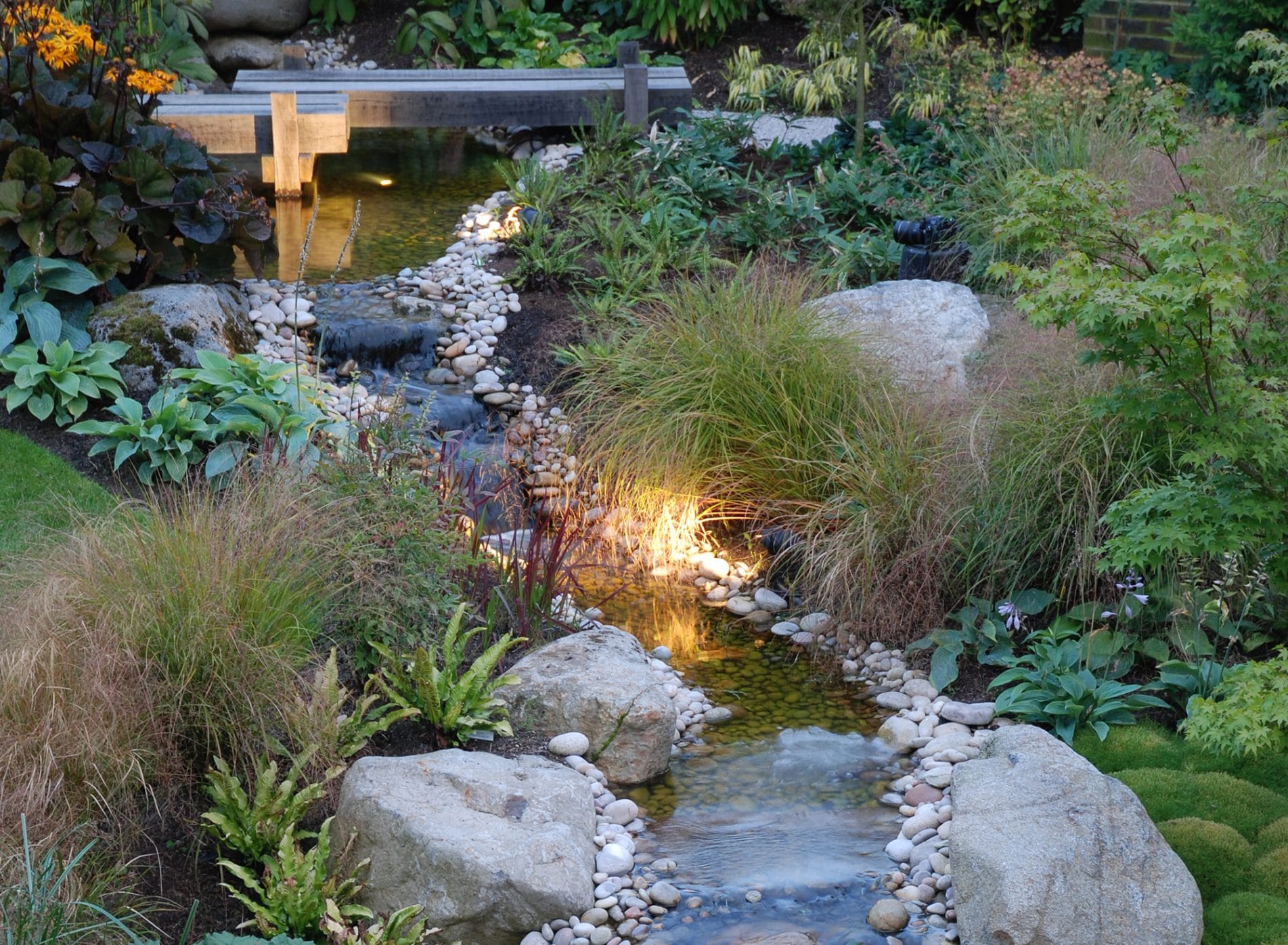 Water feature with wooden bridge and surrounded by stones, grasses and plants