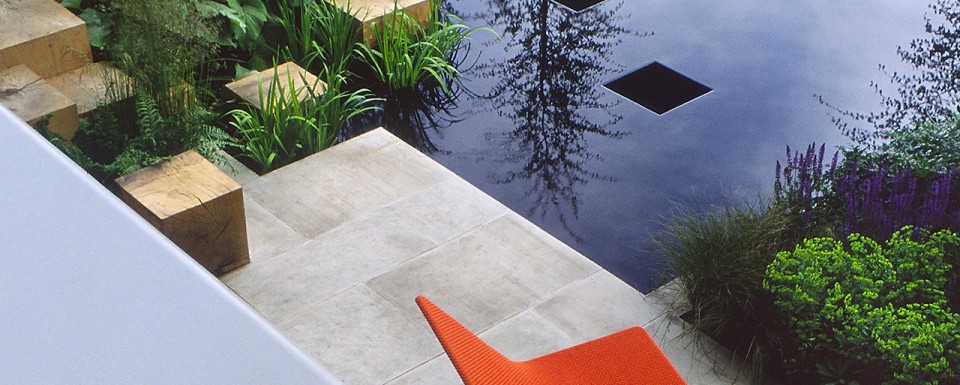 Modern Garden With Two Orange Chairs Wooden Block Rock Garden With Green Plants. Modern Water feature With Reflection Of Tree