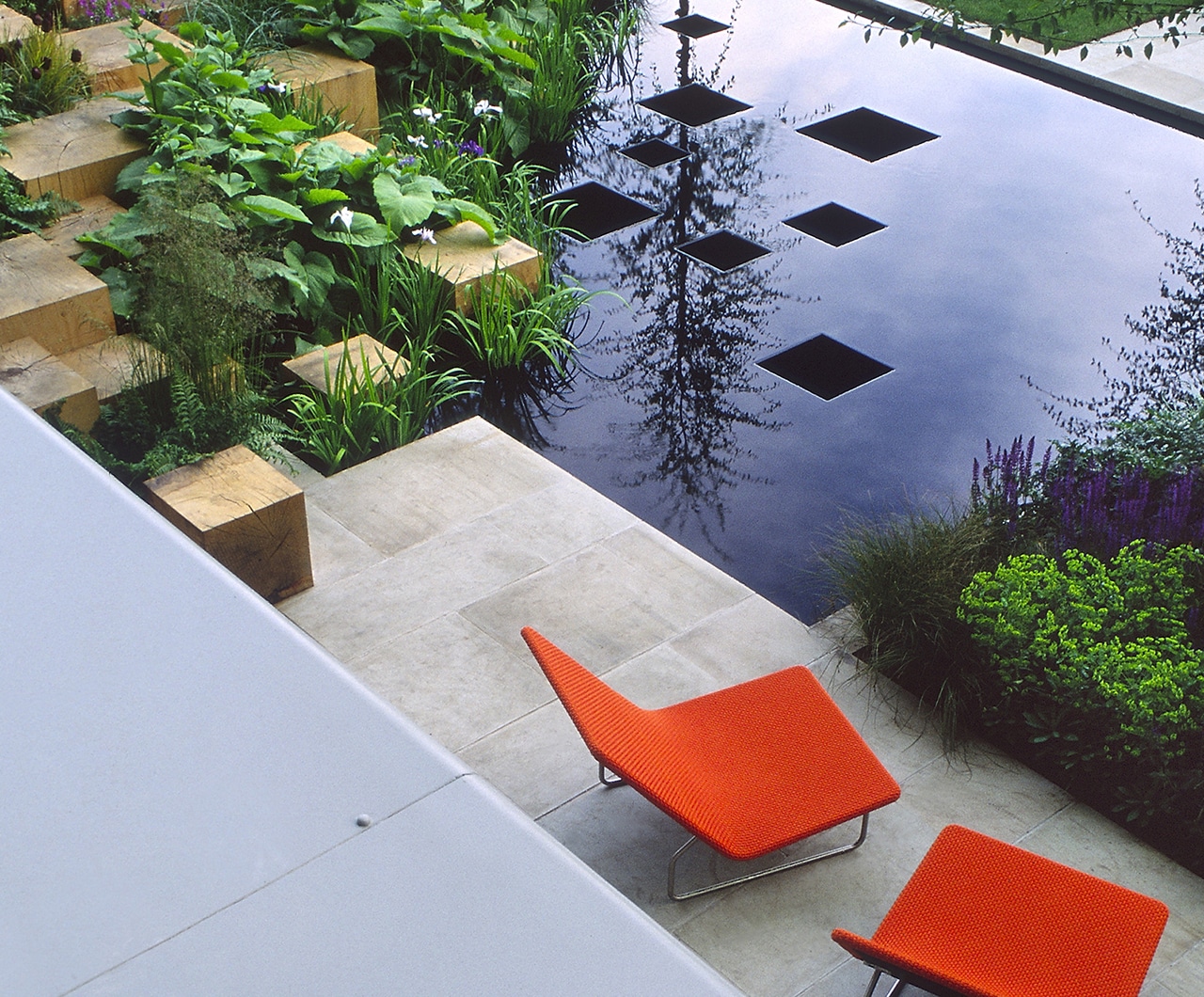 Modern Garden With Two Orange Chairs Wooden Block Rock Garden With Green Plants. Modern Water feature With Reflection Of Tree