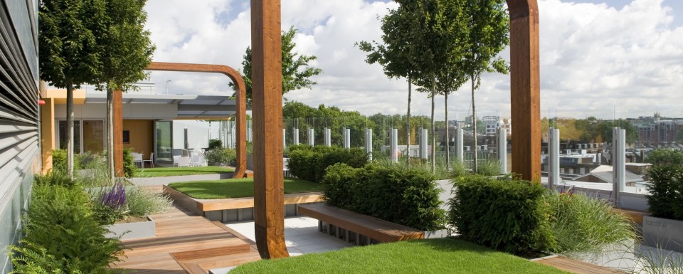 Modern City Rooftop Garden Featuring Squares Of Grass Surrounded By Wooden Decking With Seating Area And Wooden Arches