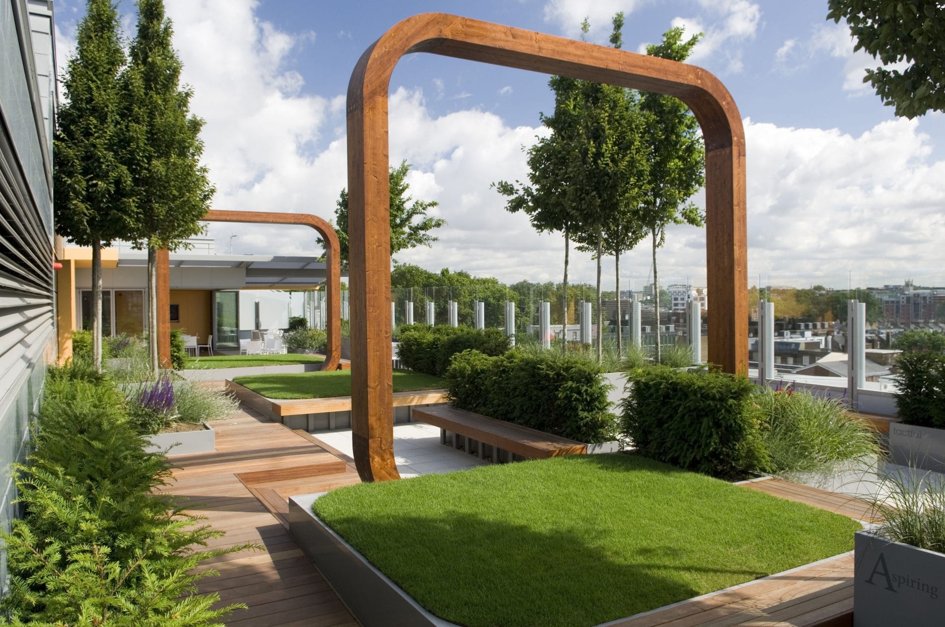 Modern City Rooftop Garden Featuring Squares Of Grass Surrounded By Wooden Decking With Seating Area And Wooden Arches